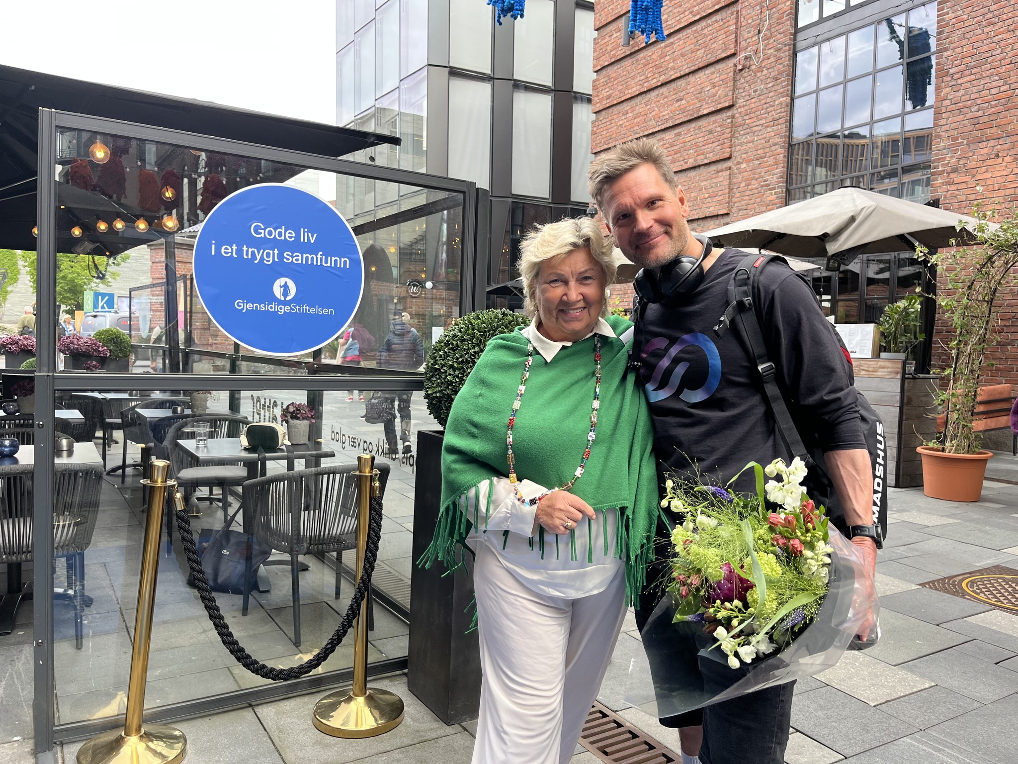 Marianne og Øystein "Pølsa" Pettersen smiler og står tett sammen foran et skilt med teksten "Gode liv i et trygt samfunn" fra Gjensidigestiftelsen. Øystein holder en blomsterbukett og har på hodetelefoner og ryggsekk.