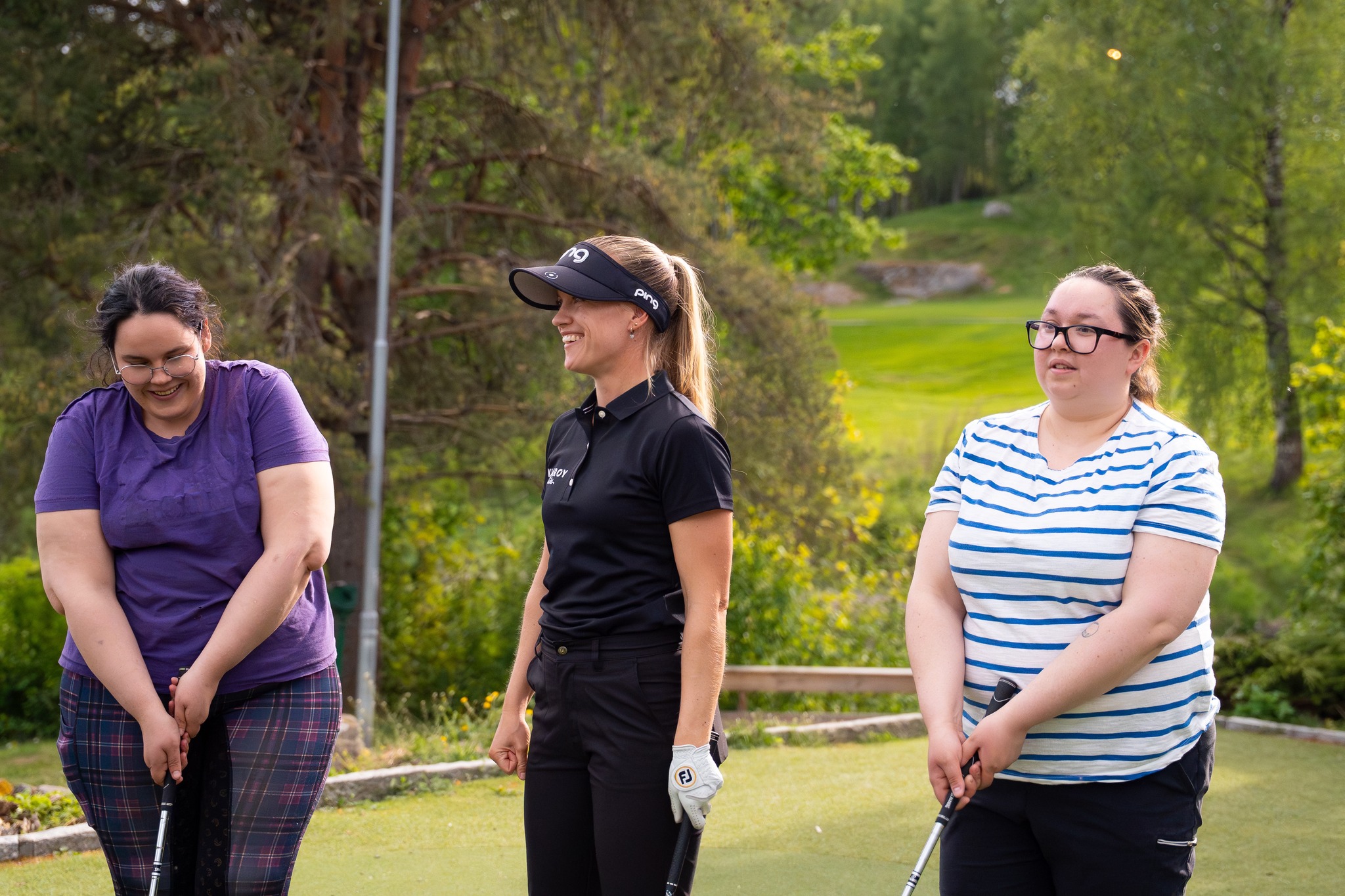 Michelle Forsland instruerer to kvinnelige deltakere i golf på en solfylt treningsøkt i grønne omgivelser.